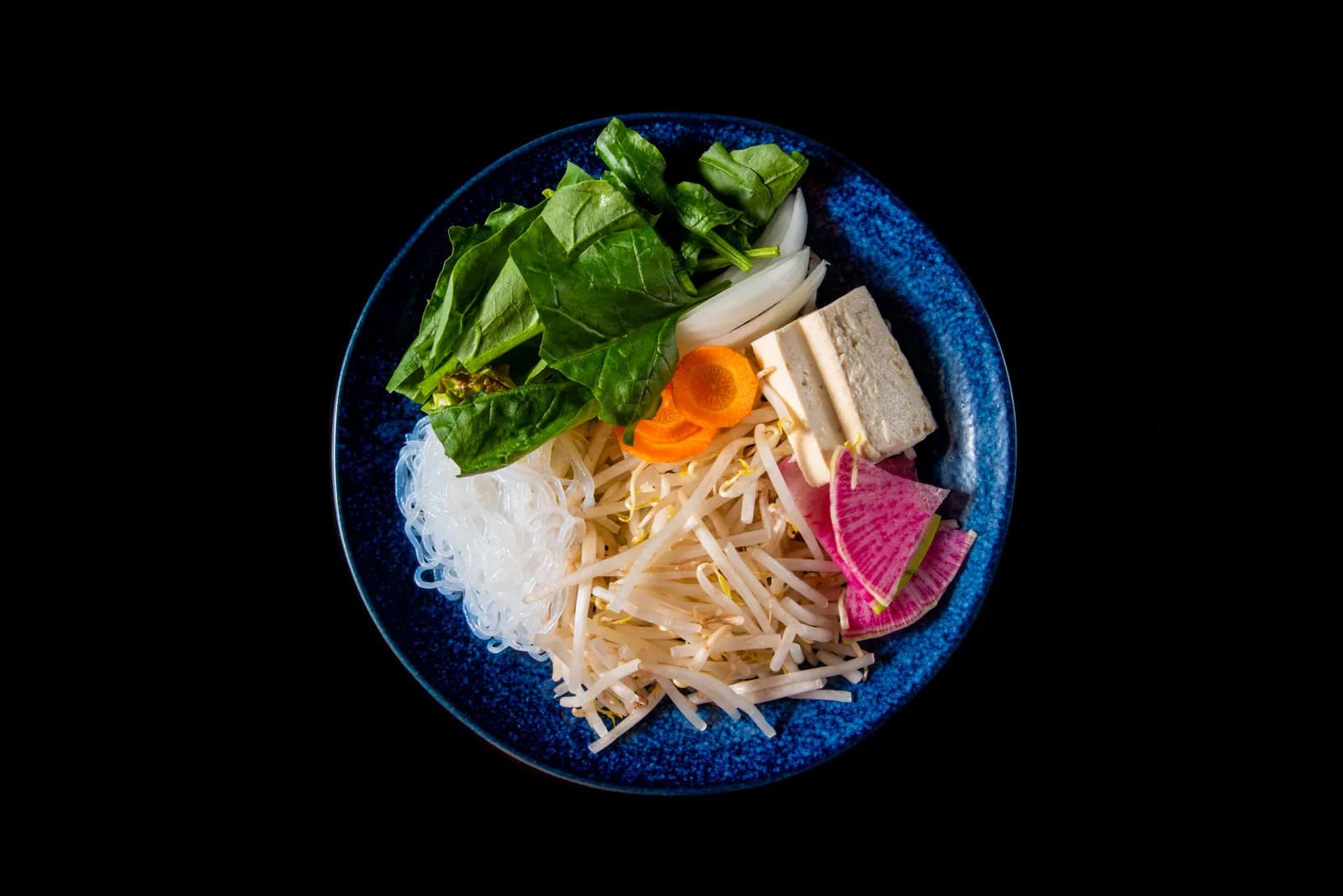 藍の平皿に盛った鍋野菜。青梗菜、豆腐、にんじん、白髪ねぎ、紅芯大根、春雨が扇形に並ぶ