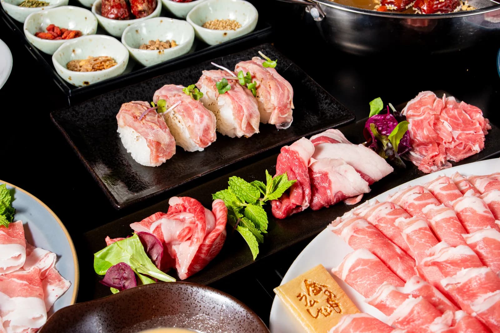 卓上を斜めから。黒い長皿の炙りラム肉寿司、スレートの肉盛り、白い器のロールラムが重なる