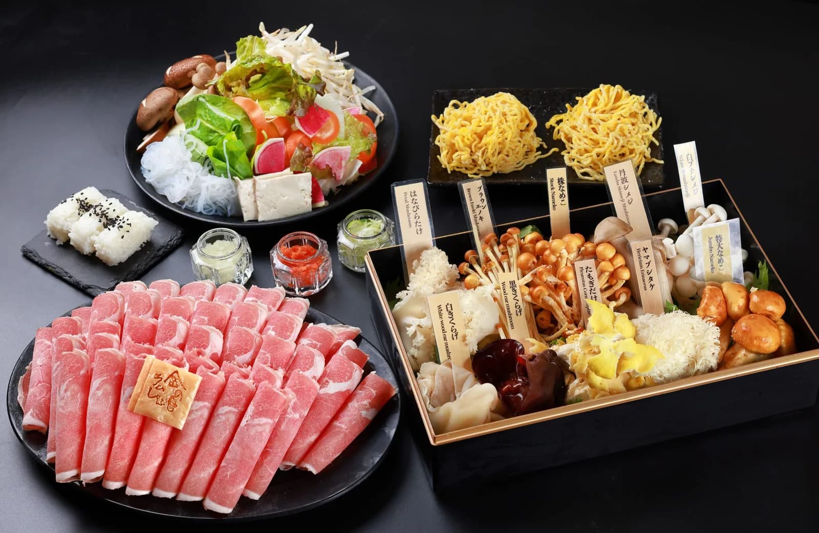 从正上方俯拍黑色餐桌。立有木牌的菌菇漆盒、薄切羊肉大盘、沙拉、两份鸡蛋面、饭团、佐料瓶一同排列