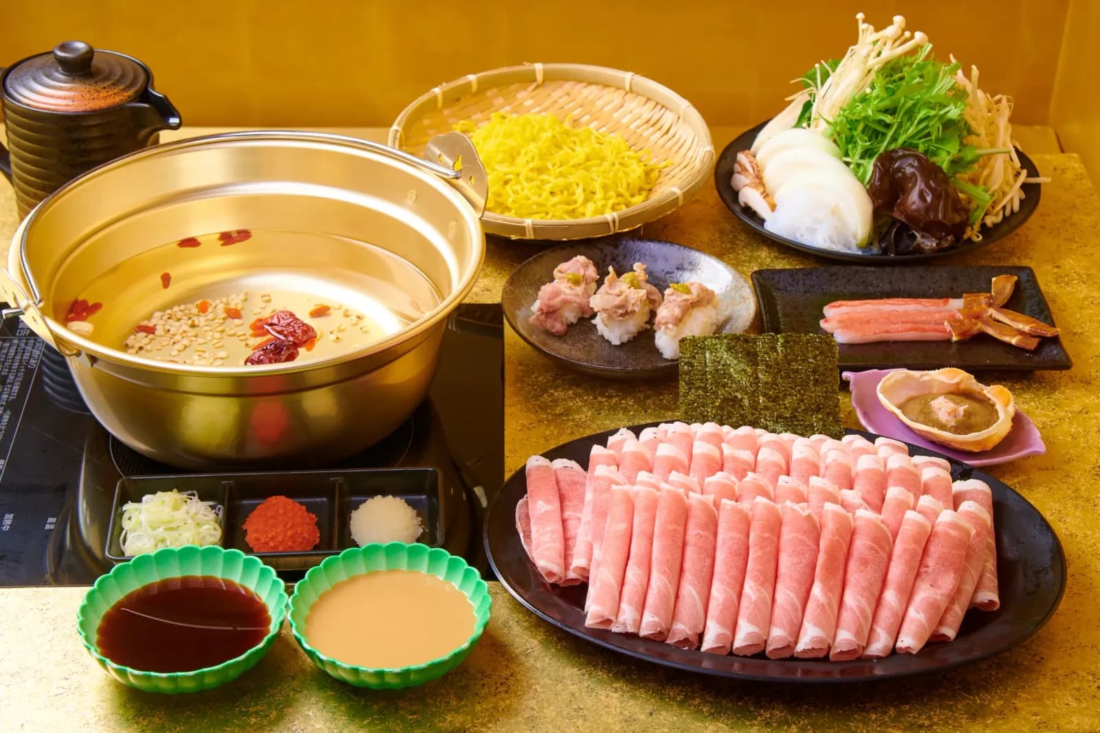 A full course spread on a gold table: a gold pot with goji berries and jujubes floating in the broth, egg noodles, sushi, crab legs, a lamb platter and two dipping sauces