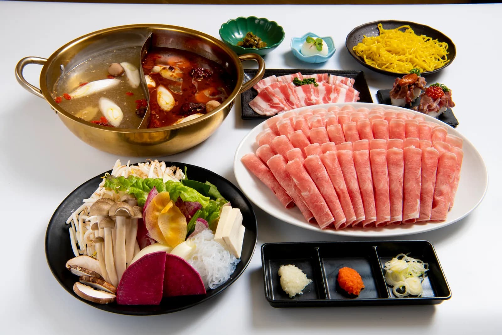 A full view of the course centered on a gold twin-broth pot, with thinly sliced lamb on a white platter alongside mushrooms, vegetables and condiments