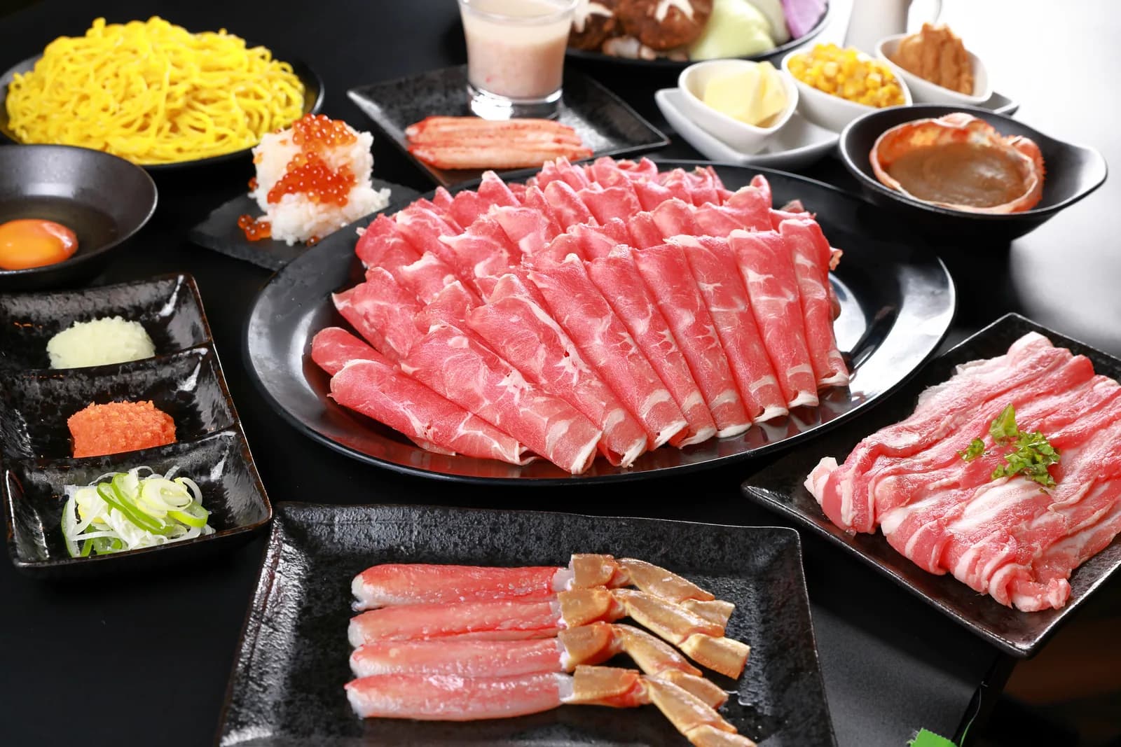 黒い地に、扇状に盛った薄切りラム肉の大皿を中心として、たまご麺・野菜・薬味・溶き卵を配したコースの全景