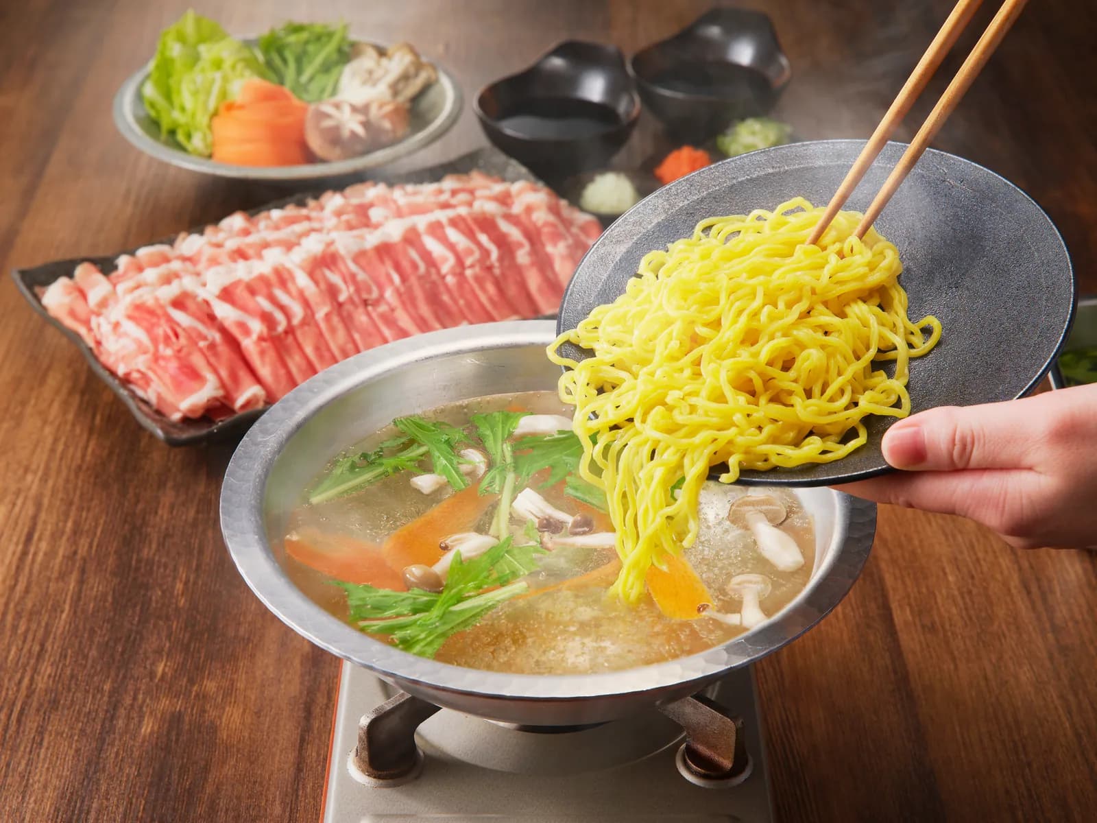 Egg noodles to finish being scooped from the pot; a platter of thinly sliced lamb and bowls of condiments sit behind