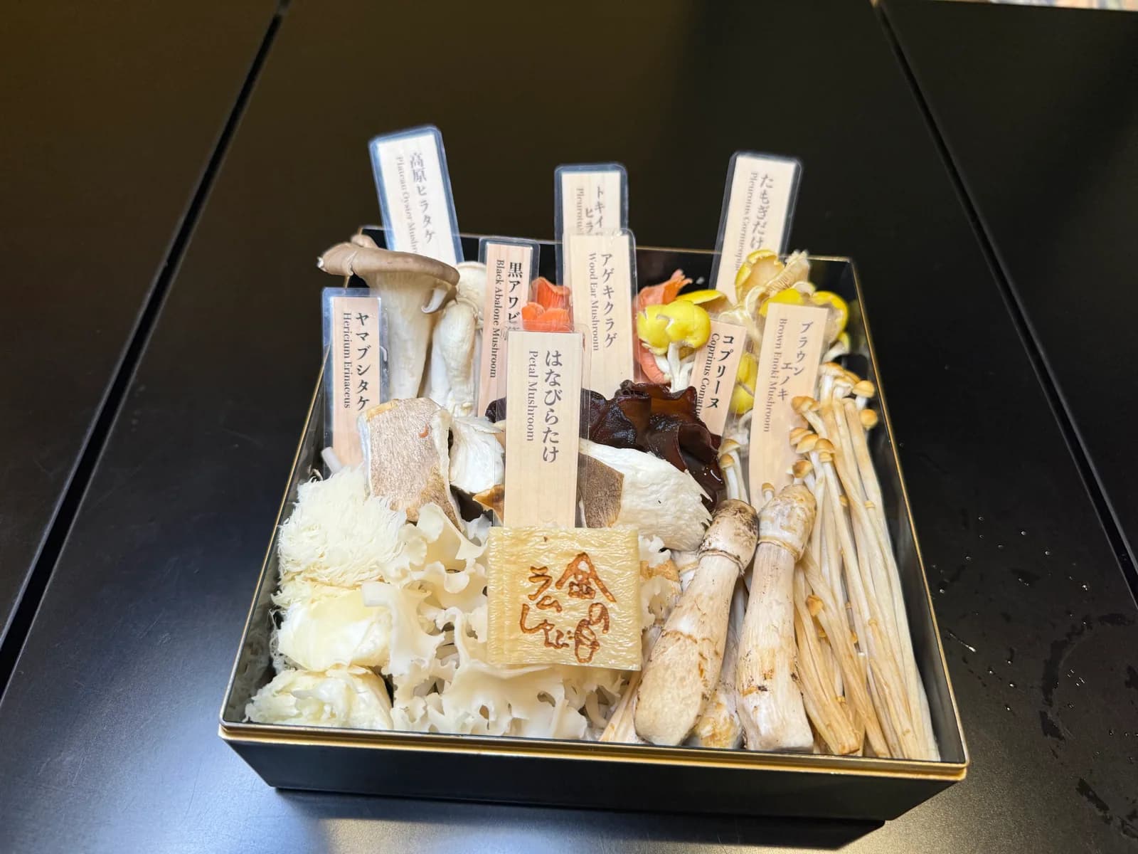 A platter of rare mushrooms: Tamogitake, Hanabiratake and others laid out in a black wooden box with variety-name tags