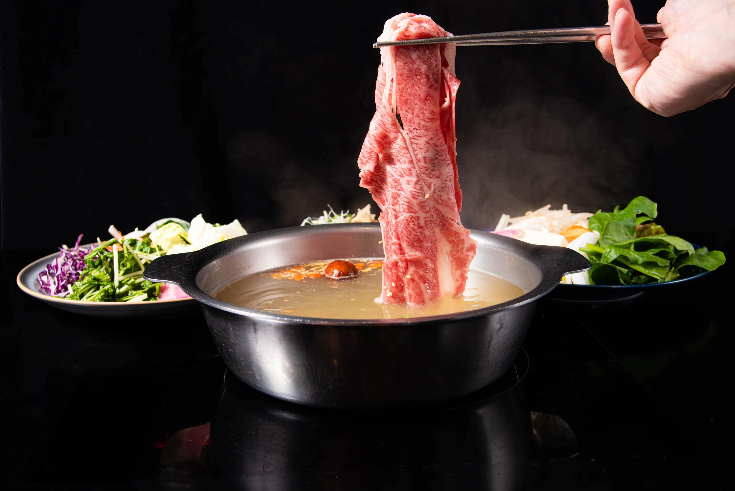 Thinly sliced lamb gripped in chopsticks and lifted high out of the paitan broth pot; steam rising, with plates of colorful vegetables lined up behind