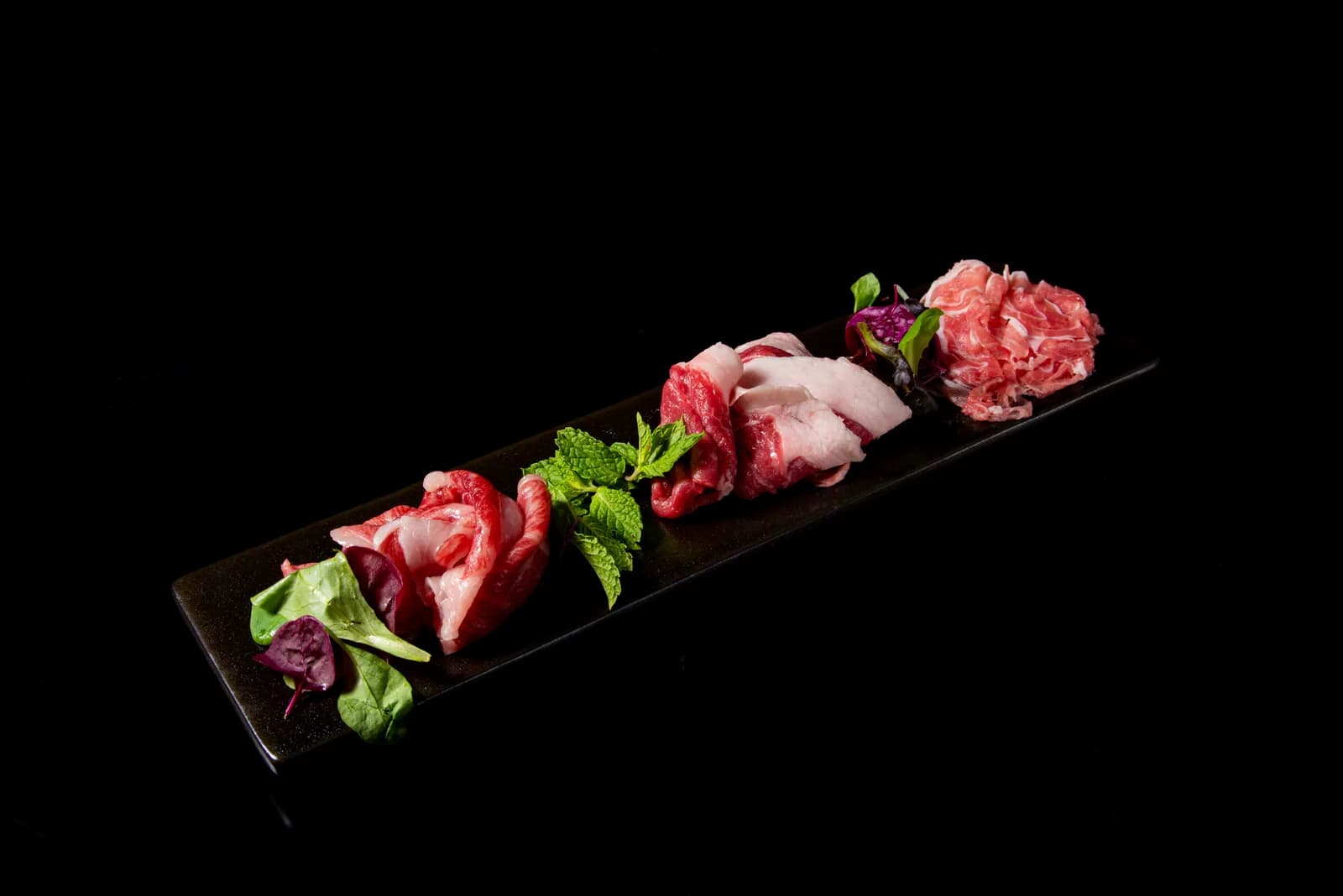 Several cuts of lamb laid out at an angle on a long black plate, garnished with mint and purple leaves, shadows falling against a black background