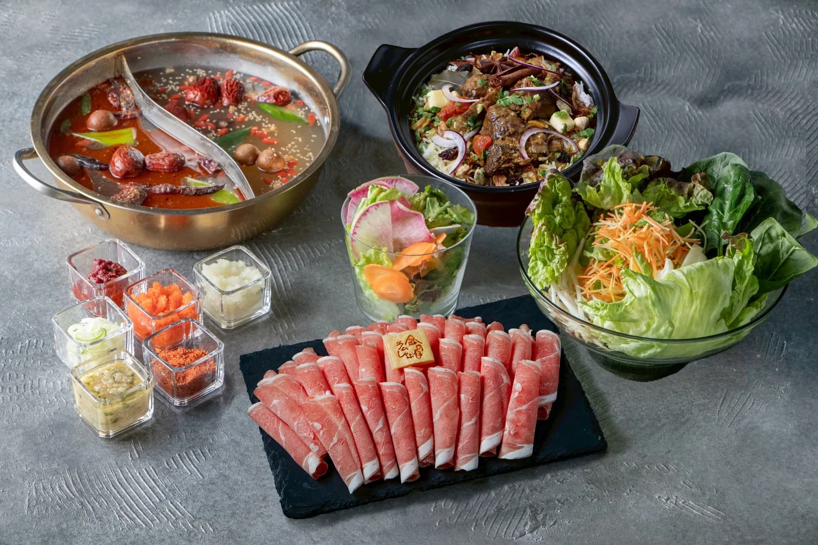 A course spread on a grey stone table: a gold twin-broth pot, stir-fried lamb in a black clay pot, salad in a glass bowl, six condiments and a lamb platter on slate