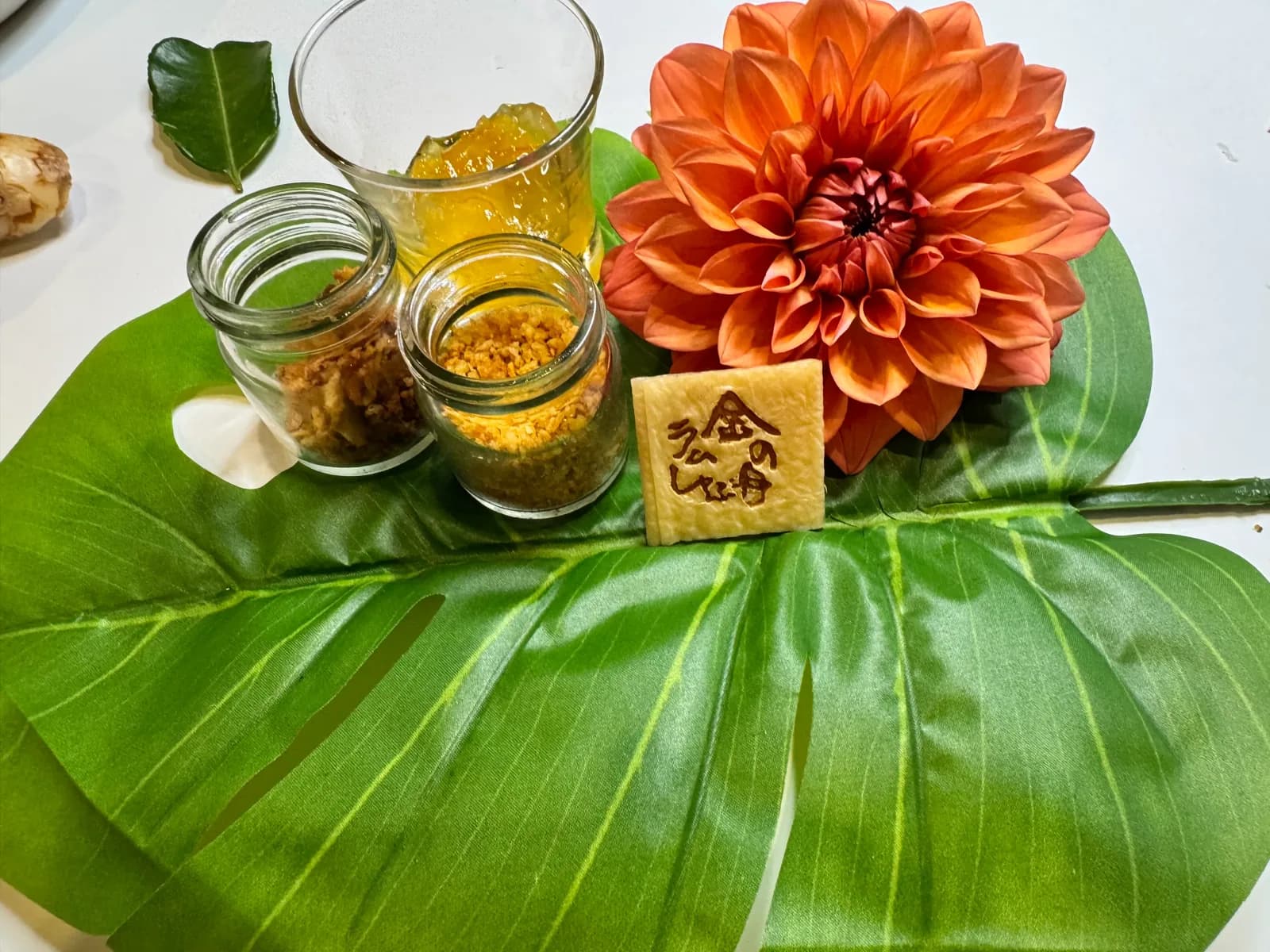 Artificial monstera and dahlias spread on a white table, three condiments including yuzu kosho in glass jars, and a KINNOME Lamb Shabu branded plate