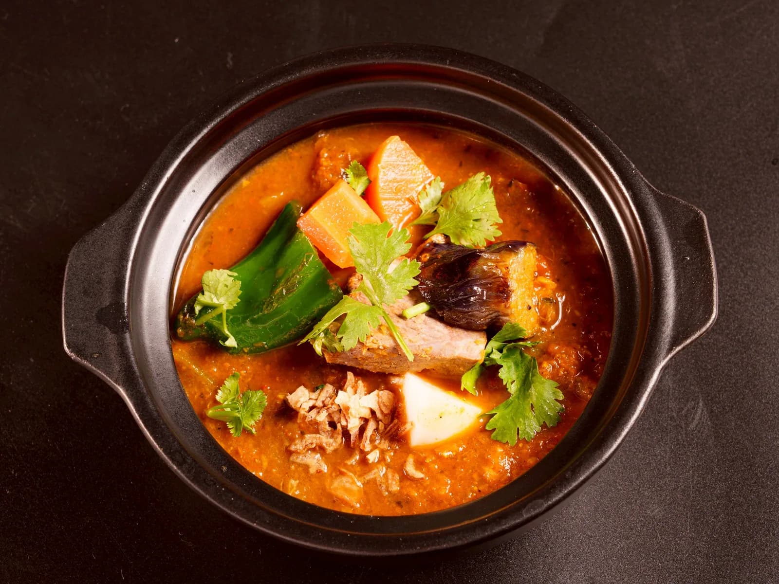 A black clay pot brimming with mala stew, seen from directly above; chunks of lamb, daikon, carrot, green pepper and cilantro float in the broth