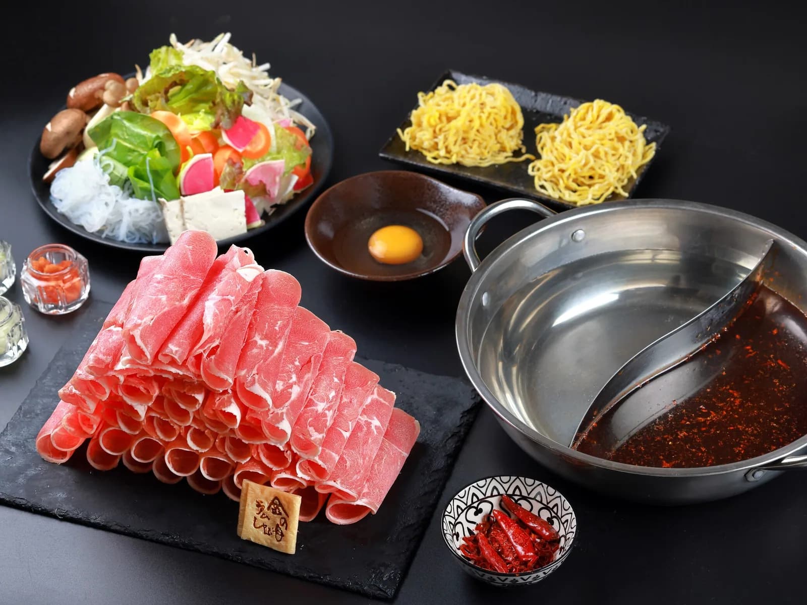 Overhead view on a black background: rolled lamb stacked in tiers on a black stone slab, a twin-broth pot of paitan and mala, two portions of egg noodles, beaten egg, a platter of vegetables and tofu, and a bowl of chili