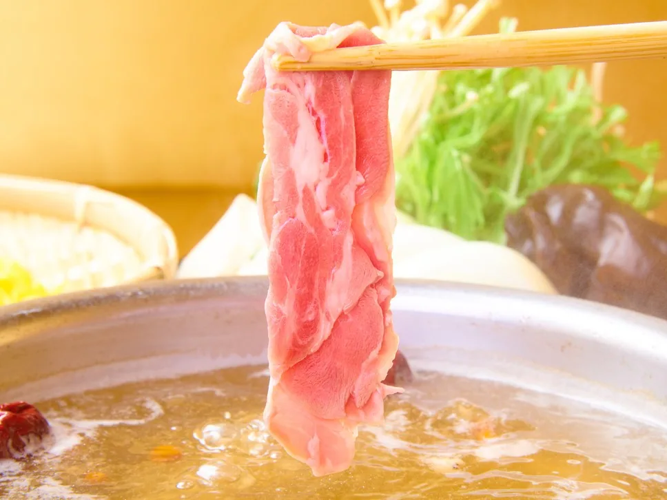 Close-up of a paitan pot against a yellow wall; bamboo chopsticks lift a single slice of lamb, with chrysanthemum greens and shiitake behind