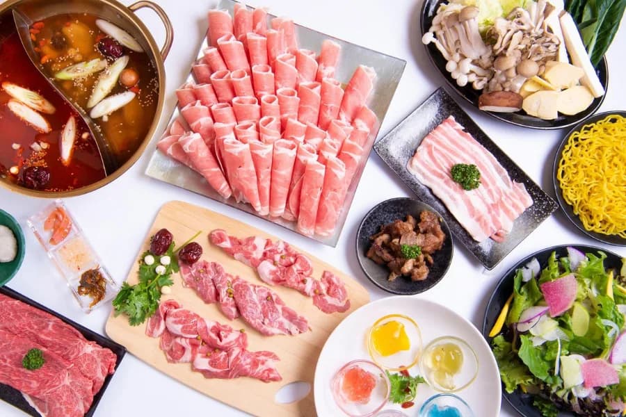 Overhead view on white: the full course with a twin-broth pot of red mala broth, a platter of rolled lamb, fresh lamb served on a cutting board, a mushroom platter, Chinese noodles, salad and small dishes of condiments