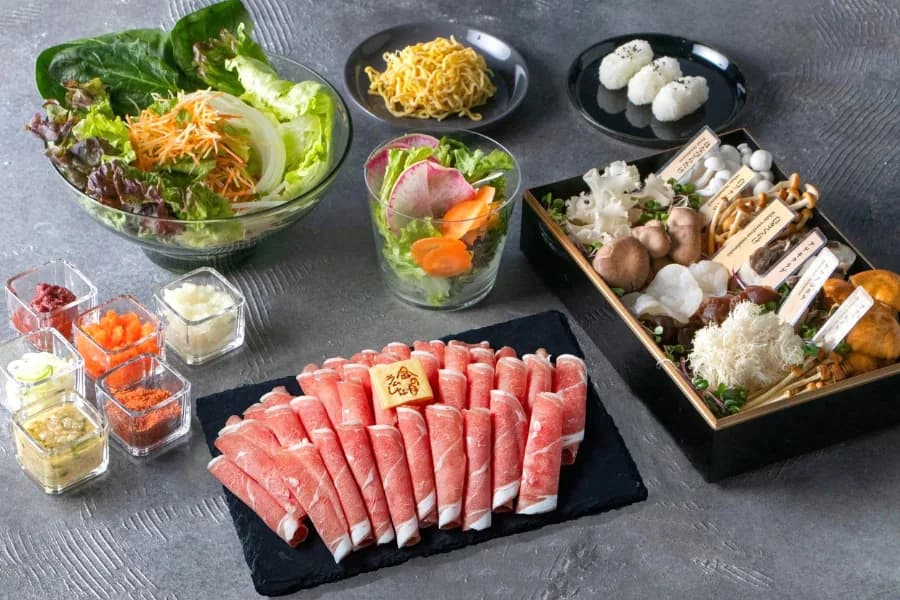 Rolled lamb fanned out on a black stone slab with a branded plate beside it, surrounded by salad, vegetable sticks, quail eggs, bowls of condiments and a lacquer box of mushrooms and side dishes, seen from above on grey