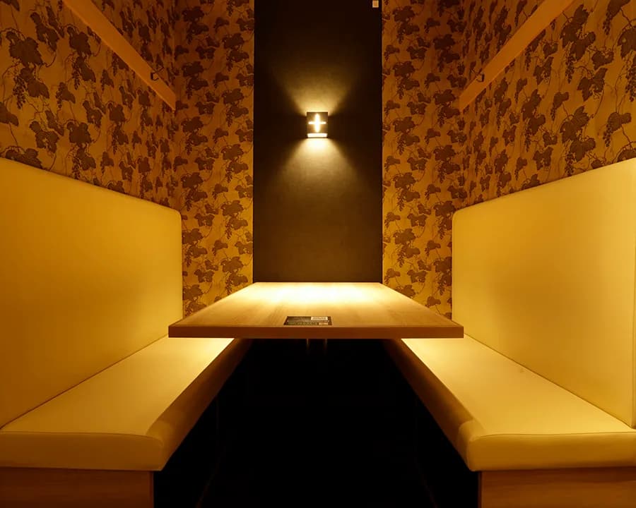 A semi-private seat at the Ikebukuro store: a wall sconce lit on the back wall between grape-patterned wallpaper, with cream bench seats flanking a wooden table
