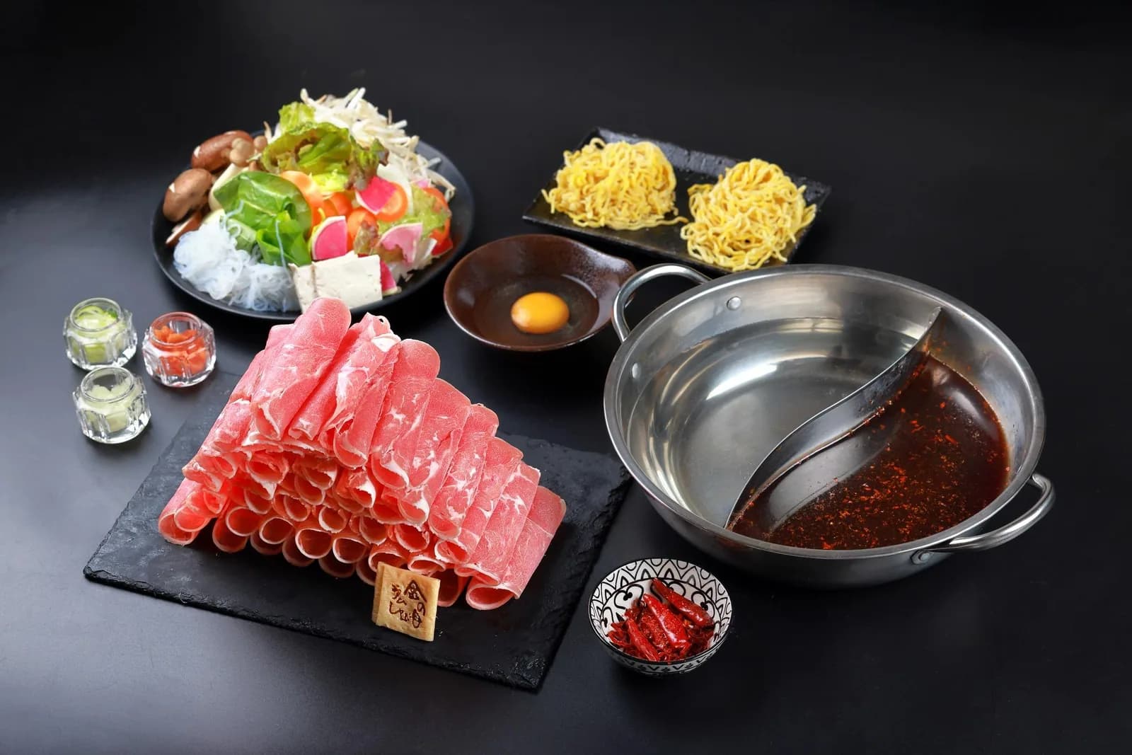 Overhead view of a tabletop on black: a twin-broth pot of mala and paitan, slate lined with rolled lamb, salad, egg noodles, raw egg and condiments