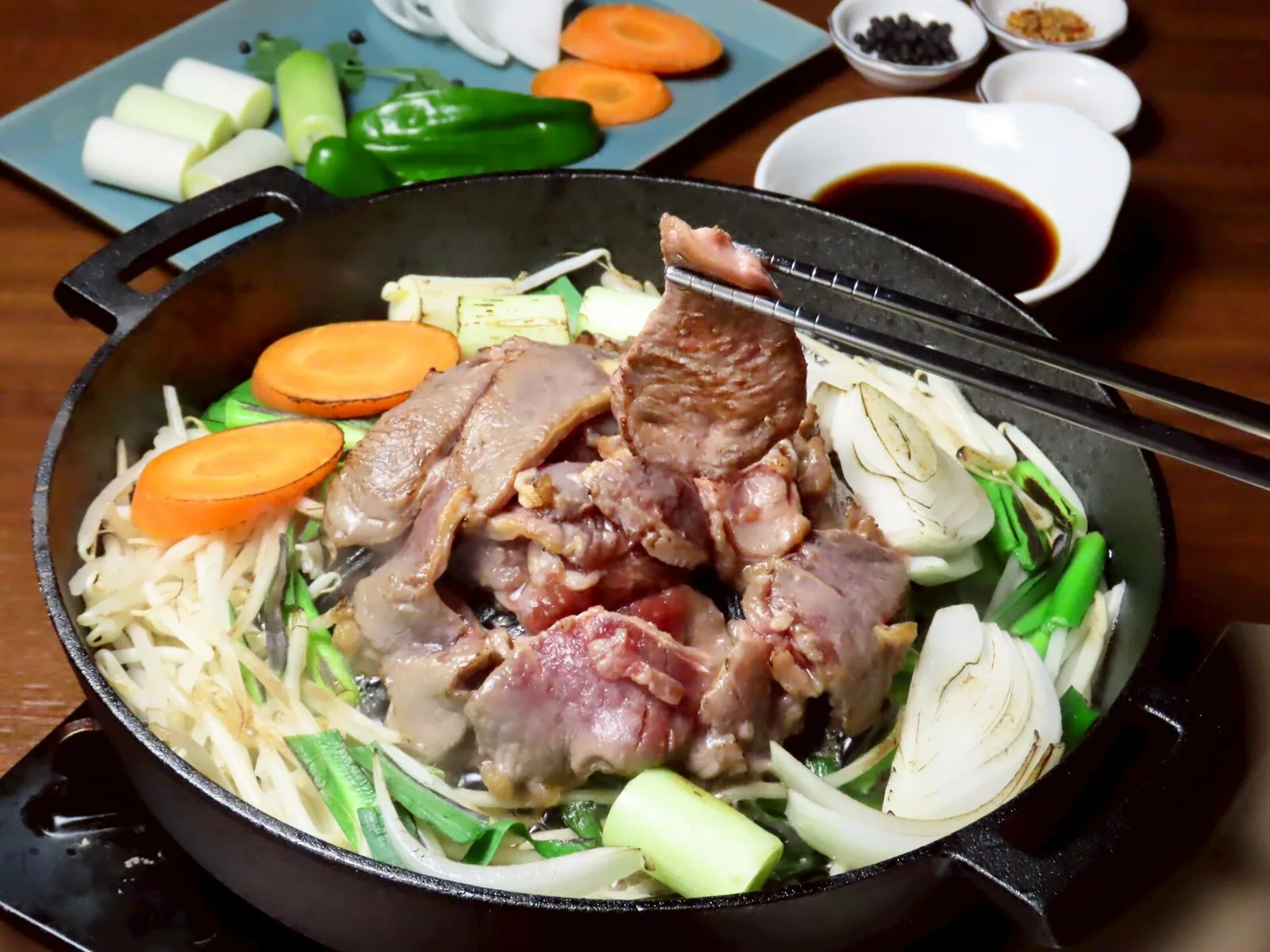 Lamb sukiyaki simmering in an iron pot; chopsticks lift browned lamb with bean sprouts, leek and carrot, with dipping sauce and condiment dishes behind