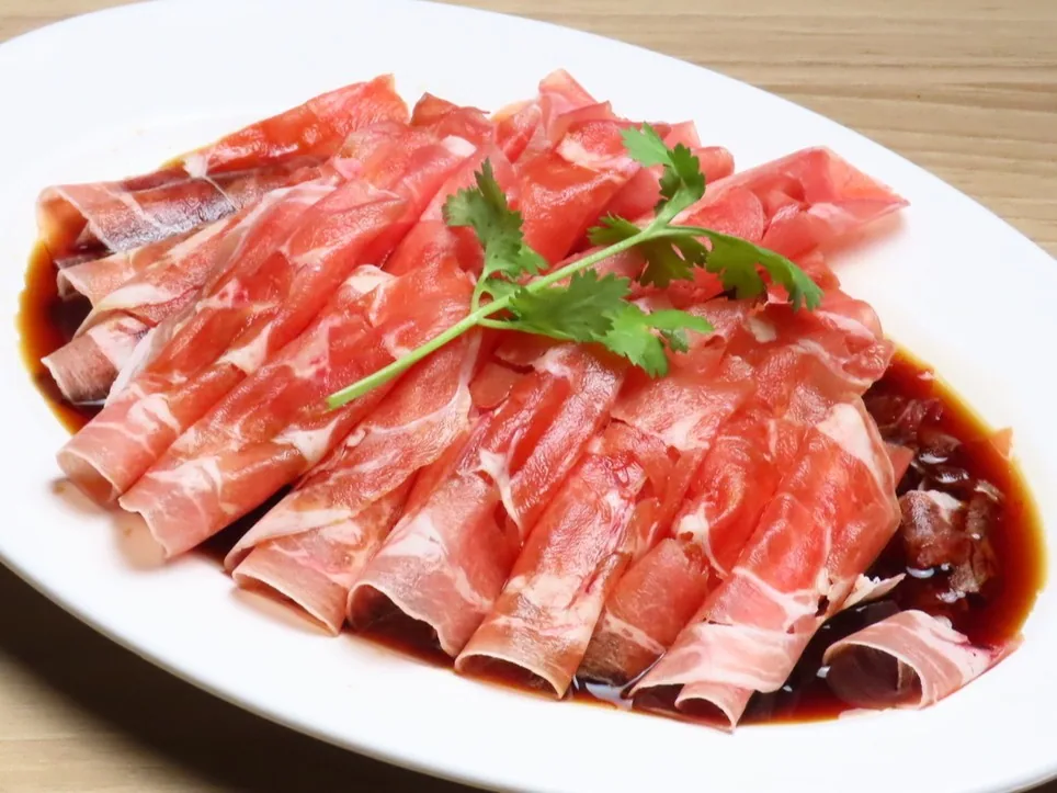 A cold dish of thinly sliced lamb mounded on a white plate, drizzled with black vinegar sauce and topped with cilantro, set on a wood-grain table