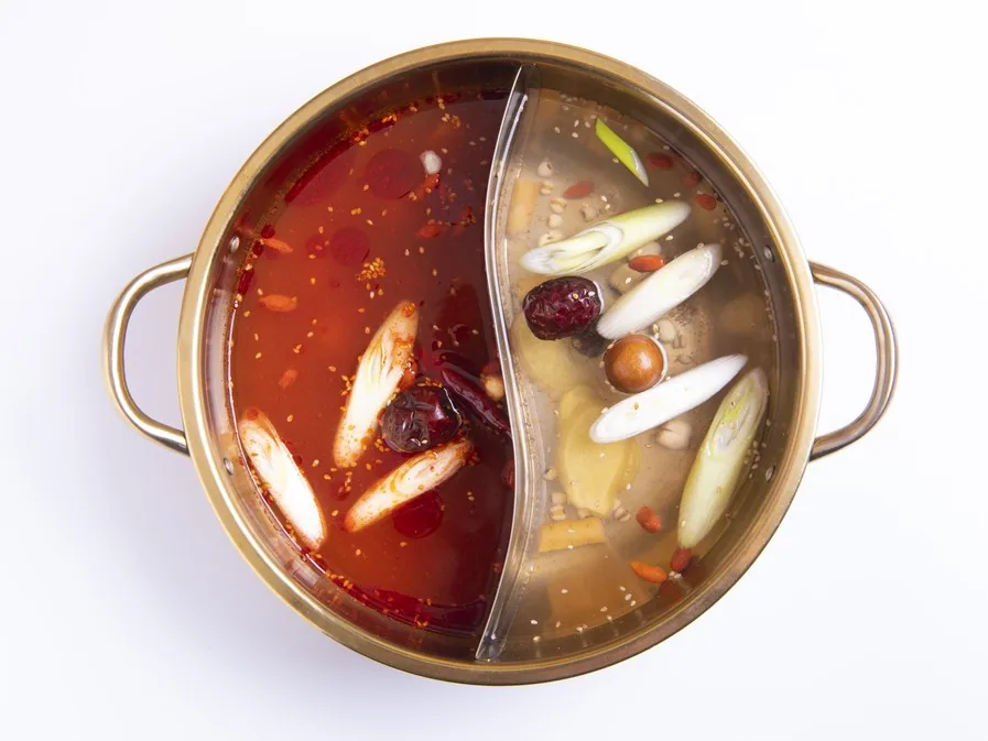A gold twin-broth pot shot from directly above on white: mala broth with floating chilies on the left, yakuzen paitan with jujubes and goji berries on the right