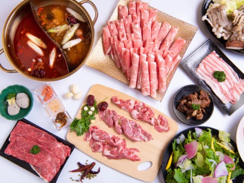 An overhead view of the full course laid out on white: a gold twin-broth pot, thinly sliced lamb, lamb steak, fresh lamb, offal, mushrooms and salad
